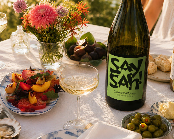 A bottle of Saysah Organic Brut on a summer table with flowers, figs, a peach salad, olives and bread
