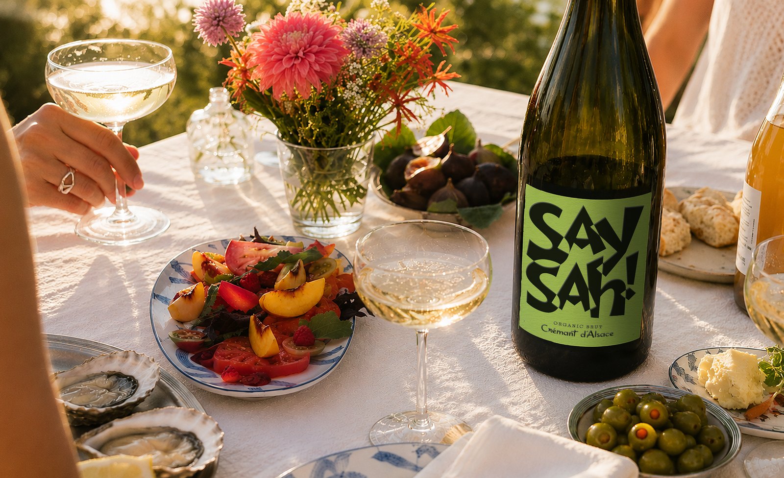 A bottle of Saysah Brut on a summer table with coupes, oysters, peaches and olives